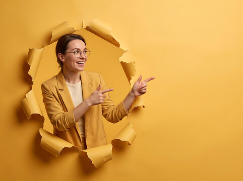 Young woman in yellow blazer pointing through torn hole in yellow background 