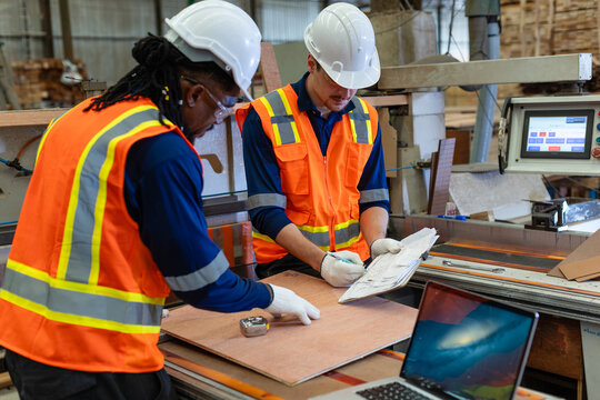 Plywood for woodworking rests beside a carpenter who touches its surface while his colleague writes on a clipboard.