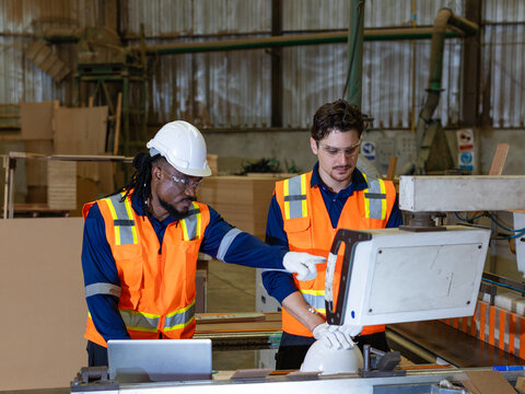 Inside a woodworking shop, a carpenter explains plywood work to his colleague, pointing at the wood workshop control panel.
