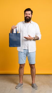 A stylish man with a beard, wearing a white shirt and blue shorts, proudly holds up a blue shopping bag. He stands against a vibrant yellow background.