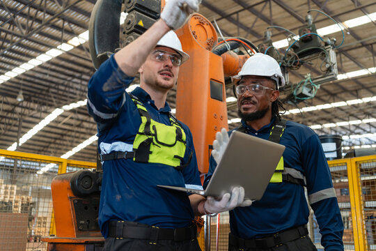 Checking industrial robot automation during inspection, a man holding a laptop points upward beside his colleague inside the fenced cell.
