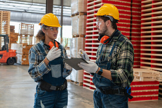 Discussing pallet logistics in the warehouse, a woman and her colleague consult a tablet beside red wooden pallet stacks.