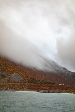 Aerial view of the Lofoten mountains featuring vibrant autumn foliage, rocky terrain, and low-hanging clouds above the sea in Vagan Municipality, Nordland, Norway.