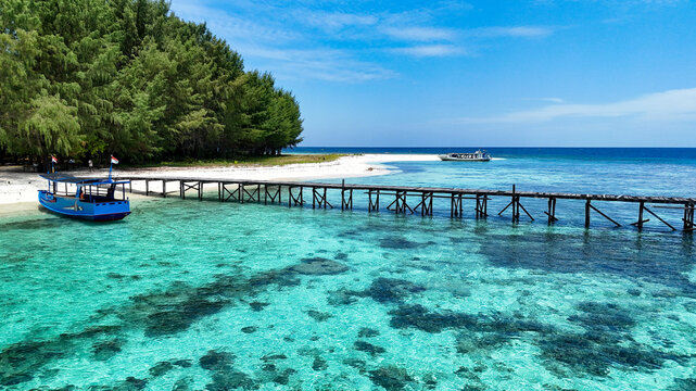 Aerial view of Pulau Cilik with a wooden pier extending into clear turquoise water, a blue boat moored nearby, and lush trees along the shore in Karimunjawa, Central Java, Indonesia.