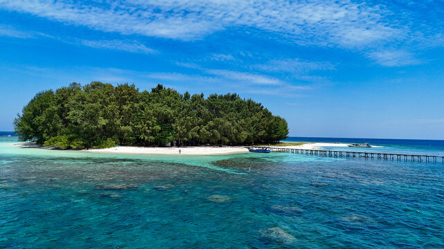 Aerial view of Pulau Cilik with lush green trees, white sandy beach, wooden pier extending into turquoise sea, and clear blue sky in Karimunjawa, Central Java, Indonesia.
