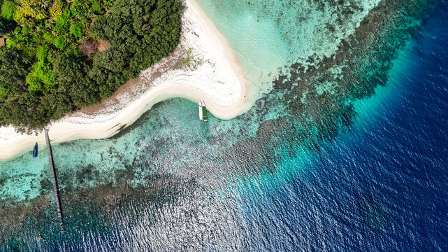 Aerial view of Pulau Cilik with white sand beach, lush vegetation, wooden pier, and boats in clear turquoise waters over a coral reef in Karimunjawa, Central Java, Indonesia.
