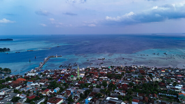 Aerial view of the mosque, dense residential rooftops, a long pier extending into the turquoise sea, and calm coastal waters in Karimunjawa, Central Java, Indonesia.