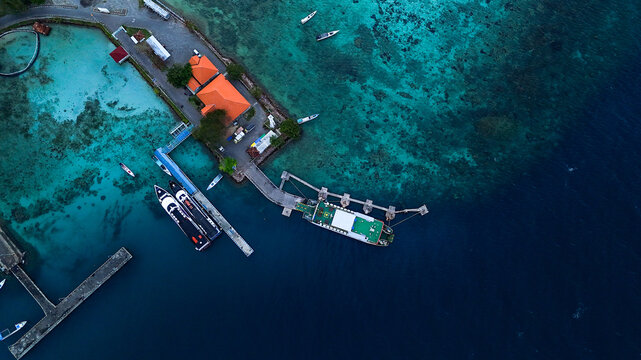 Aerial view of Port Karimunjawa featuring a pier with docked passenger boats, turquoise waters, and buildings with orange roofs in Karimunjawa, Central Java, Indonesia.