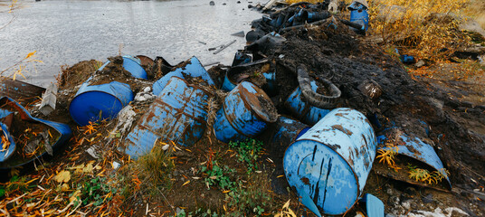 Blue barrels and dark oily debris scattered along the shore beside calm water, showing an environmental pollution scene with industrial waste and autumn vegetation © Кирилл Горшков