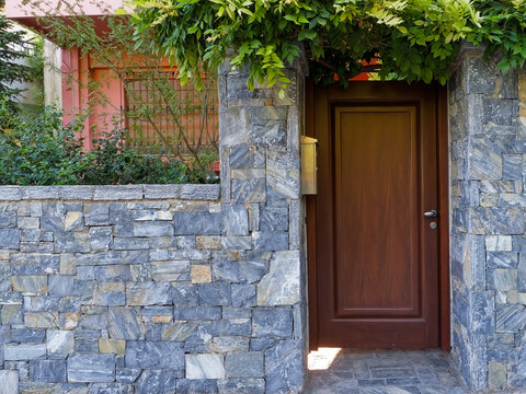 Exterior view of a private wooden entrance door set within a stone boundary wall with lush foliage in Athens, Greece.