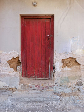 Vertical view of a locked old red wooden door set in a crumbling stucco wall with exposed clay masonry in Athens, Greece.