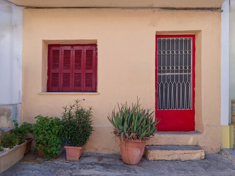 Front view of a humble traditional Greek house with crimson red wooden shutters, door, and potted plants in Athens, Greece.