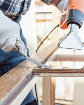 Adult carpenter wearing protective leather gloves, using a heat gun and a wood scraper to remove paint from an old wooden window. Construction industry, carpentry. Restoration. 