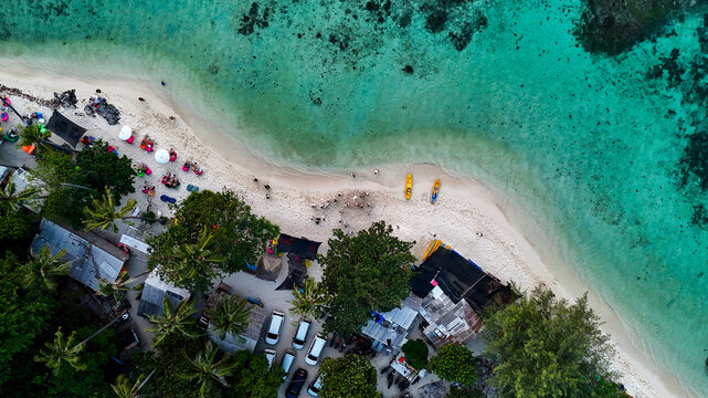 Aerial view of Pantai Tanjung Gelam with white sand beach, turquoise water, lush palm trees, small buildings, and parked cars in Karimunjawa, Central Java, Indonesia.