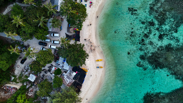 Aerial view of Pantai Tanjung Gelam featuring a white sandy beach, turquoise coral reef waters, lush palm trees, and parked vehicles near the shore in Karimunjawa, Central Java, Indonesia.