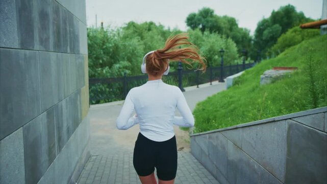 Woman jogs uphill with headphones on. Her hair flows with motion. Gray stone walls flank the path. Greenery and lamps line the route. Active lifestyle captured in motion. Great for fitness, health