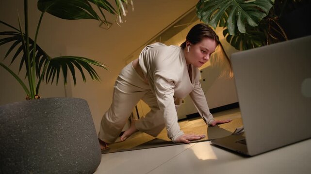 Woman practicing indoor yoga stretches on mat among tropical plants with warm light and laptop close