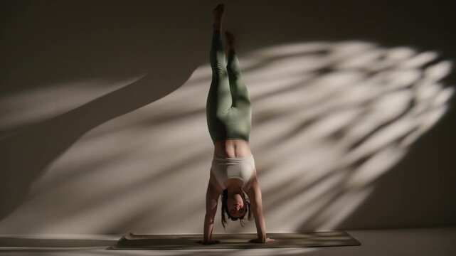 Handstand yoga pose in dim studio with diagonal light, balanced inversion on yoga mat, calm balance.