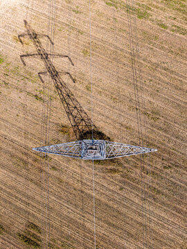 Aerial view of a high-voltage power line pylon casting a long shadow over a harvested agricultural field in Brasov, Brasov, Romania.