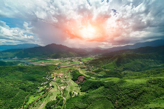 Aerial landscape of green mountains, winding river, agricultural fields and a village beneath dramatic sunset clouds in central Vietnam, viewed from above.