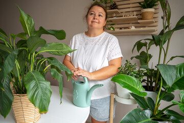 A woman stands among lush houseplants in a bright indoor setting, holding a watering can. The scene illustrates home gardening, urban jungle lifestyle, and personal wellness through nature. © JULIA