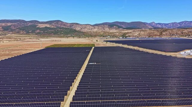 Rows of solar panels cover a large field in a rural location. Mountains rise in the distance under a clear blue sky. The area shows no signs of buildings or people.