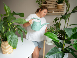 A middle-aged woman waters lush green indoor plants with a watering can in a bright home environment. This image represents domestic wellness, urban gardening, and a sustainable modern lifestyle. © JULIA