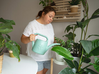 A woman waters indoor potted plants with a green watering can in a bright room, representing home gardening, plant care, and a sustainable lifestyle focused on wellness and nature. © JULIA