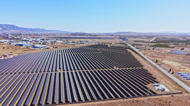 A solar farm covers a vast area of land in the countryside with many solar panels aligned in rows. The scene shows a clear sky and distant mountains.
