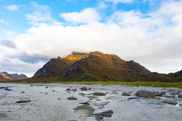 View of Lofoten mountains rising above a rocky beach with green grass and scattered stones under a cloudy sky in Vagan Municipality, Nordland, Norway. © Travel 'n' Lifestyle