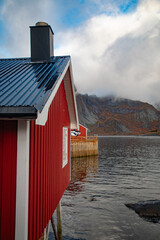 View of a traditional red wooden cabin on stilts over the water in the Lofoten islands, with rugged mountains and a cloudy sky in the background in Vagan Municipality, Nordland, Norway. © Travel 'n' Lifestyle