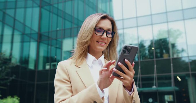 Businesswoman confidently using smartphone outside modern glass office building, daytime urban scene, smiling and tapping screen in professional interaction with device