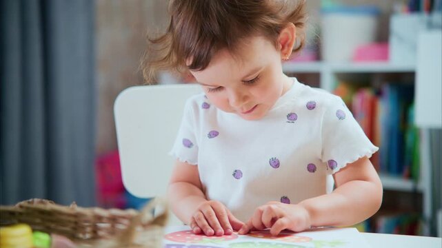 Tiny child sits at table, coloring with crayons. Brown curls frame face as she concentrates. Soft focus background holds colorful toys and books. Bright room offers warm, playful, cozy atmosphere