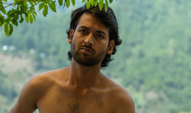 A good looking Indian bearded shirtless man with a serious expression looks directly at the camera with his head tilted under green leaves against a blurred background.