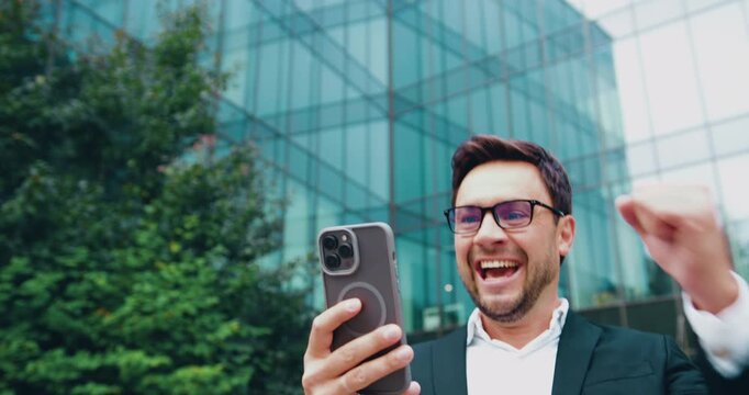 Attractive young man wearing white shirt holding phone and celebrating success, looking at cellphone screen, raising fist with excitement at modern glass building background outdoors