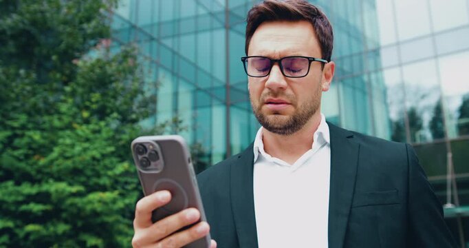 Portrait of a worried businessman holding a smartphone and looking at the screen outdoors in a city. Concept of stress, urgent message, business communication, and mobile technology.