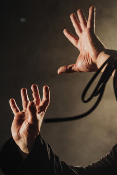 Two raised adult hands reaching upward in dramatic warm side light against dark smoky background and looping cord near wrist, palms and fingers spread showing