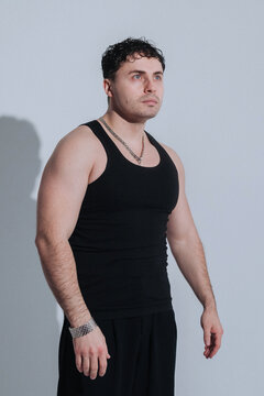 Young man in black tank top with silver chain necklace looking upward in studio portrait and strong shadow lighting that emphasizes muscular arms and focused
