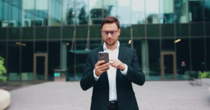 Portrait of serious and successful Caucasian businessman in suit and glasses, standing on the street in front of modern office building, confidently looking at the camera headshot of corporate manager