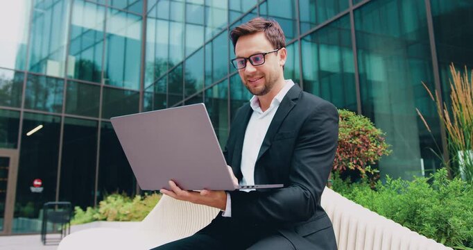 Young businessman sitting on outdoor bench in modern plaza using laptop with confident smile and daylight reflection on glass building windows, sideview portrait showing typing gesture and relaxed