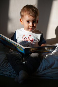 Toddler boy sitting on bed reading picture book in warm sunlight with curious expression and soft bedroom setting for parenting and early learning imagery -