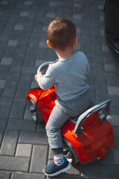 Toddler sitting on red ride-on toy car on paved sidewalk captured from behind in natural light to show playful outdoor activity and casual clothing scene before