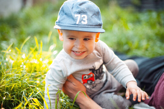 Smiling toddler boy in cap sitting on grass with adult support during warm sunlight and bokeh background that highlights joyful outdoor family moment between