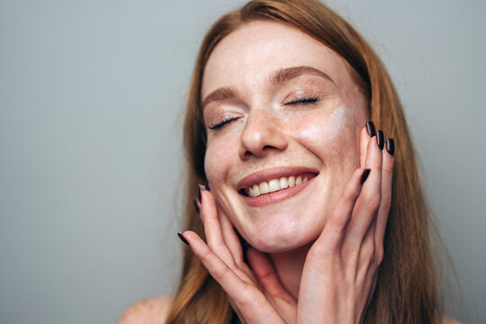 Smiling young woman with freckled skin eyes closed gently touching cheeks with manicured nails, natural beauty portrait, relaxed joyful expression, neutral