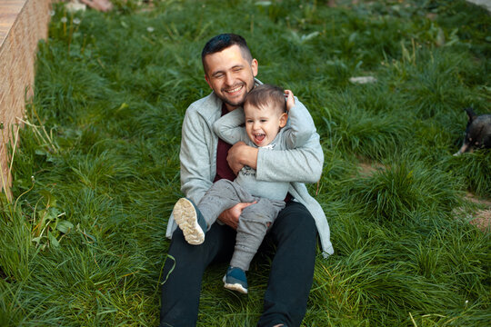 Father and toddler sitting on grass laughing and playing together outdoors in backyard setting, joyful family moment, natural light and casual clothing, closeup