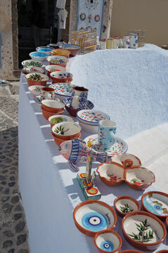 View of hand-painted ceramic bowls, mugs, and dishes with fish, olive, and evil eye patterns displayed on a white curved wall in Fira, Greece.
