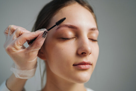 Young woman receiving eyebrow grooming with spoolie brush held by gloved hand, close-up portrait showing relaxed expression and soft studio lighting, cosmetic
