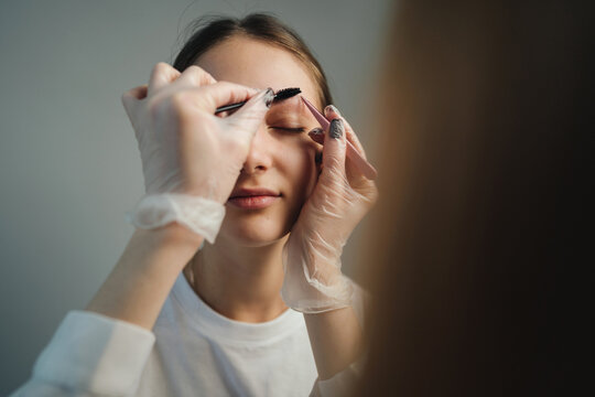 Young woman receiving eyebrow grooming from gloved technician with brush and tweezers in studio setting, eyes closed, relaxed expression and soft lighting