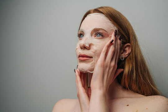 Young woman applying sheet face mask in soft studio light for skincare and self-care routine emphasis on hydration and relaxation, closeup portrait showing mask