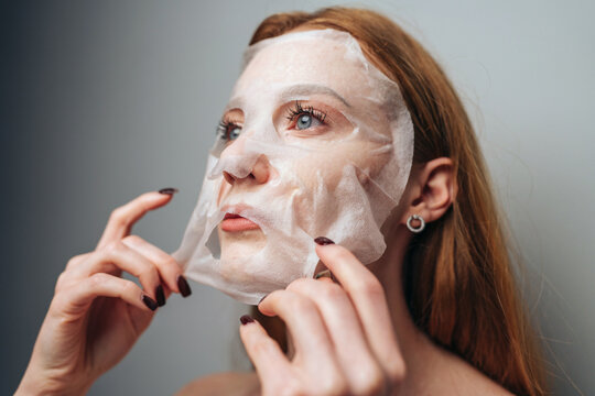 Young woman applying sheet face mask with upward gaze in soft studio lighting, skincare routine closeup for beauty and wellness themes that highlights hands